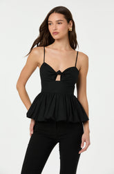 A woman is wearing a black sleeveless top with thin spaghetti straps. The top features a bustier style with a knot detail and a small cutout below the knot. It has a gathered, ruffled peplum hem that flares out at the waist.